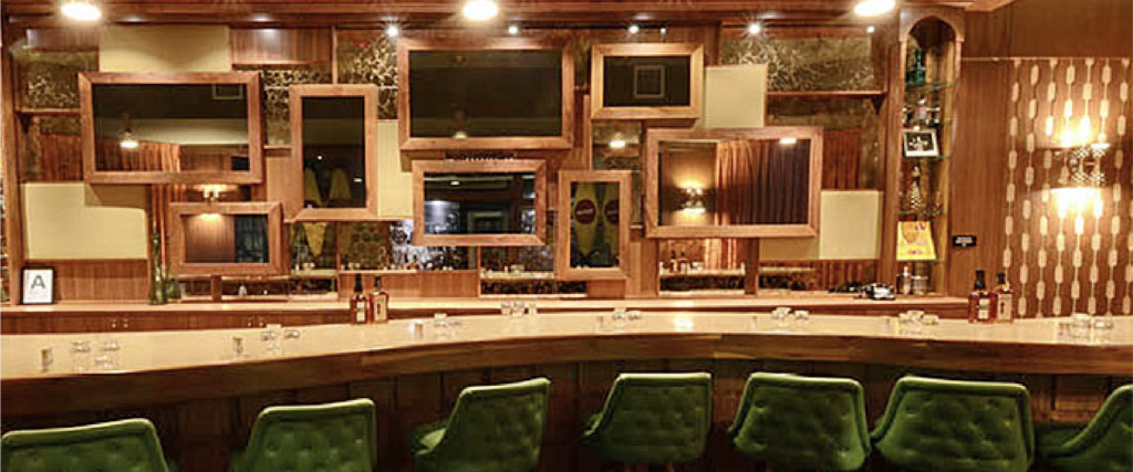 a curved bar with green chairs in front of it. Behind the bar are framed mirrors. 